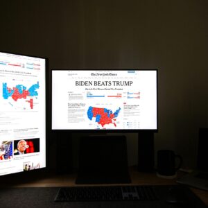 Dual computer screens in a dark room display election results indicating Biden's victory over Trump.